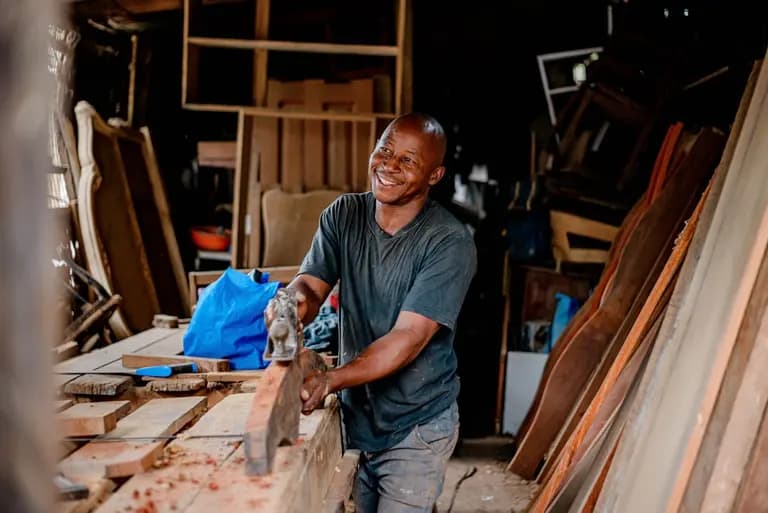 Un artisan souriant travaille le bois dans un atelier, entouré de matériaux en bois, avec une ambiance chaleureuse.