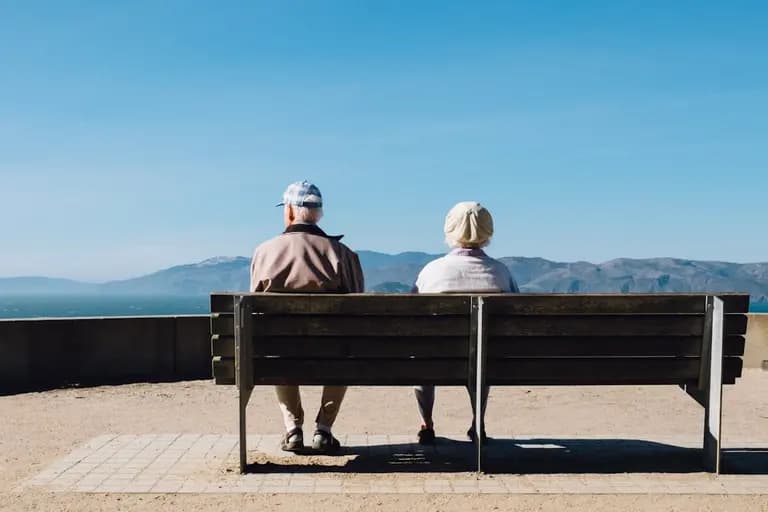 Un couple âgé assis sur un banc, regardant le paysage avec des montagnes à l'horizon sous un ciel dégagé. Ambiance calme et paisible.