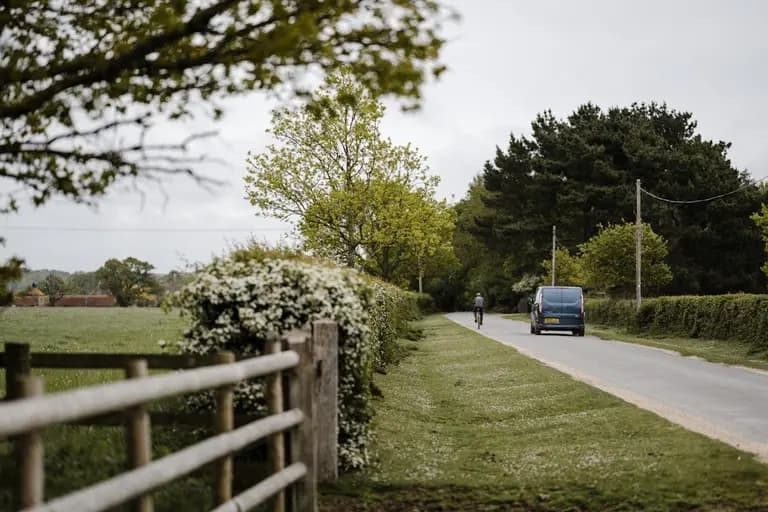 Une route tranquille bordée d'arbres et de fleurs, avec un véhicule passant à proximité, évoquant la sérénité de la campagne.